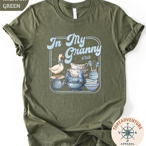 A green t-shirt with a graphic design depicting a white duck surrounded by various kitchen items, including a teapot, bowls, and a spoon.