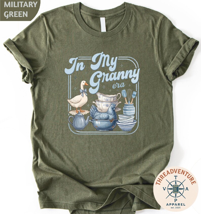 A green t-shirt with a graphic design depicting a white duck surrounded by various kitchen items, including a teapot, bowls, and a spoon.