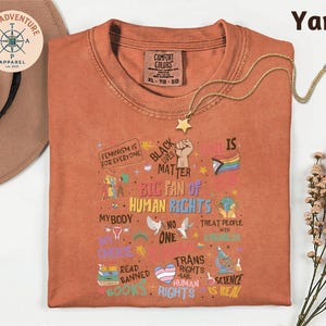 a red t-shirt with a variety of colorful text and illustrations related to human rights and social justice, including phrases like "Feminism is for everyone", "Black matter", "My body", "My choice", "Trans rights", "Treat people with kindness", "Read banned", "Books", and "Science". The t-shirt is laid out on a white surface, with a hat and some flowers visible in the background.