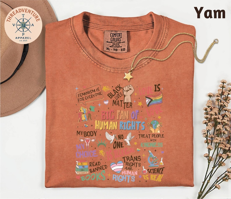 a red t-shirt with a variety of colorful text and illustrations related to human rights and social justice, including phrases like "Feminism is for everyone", "Black matter", "My body", "My choice", "Trans rights", "Treat people with kindness", "Read banned", "Books", and "Science". The t-shirt is laid out on a white surface, with a hat and some flowers visible in the background.