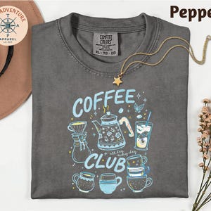 a gray t-shirt with a graphic design of coffee-related items, such as a coffee pot, a cup, and a mug, along with the text "COFFEE CLUB" printed on it. The t-shirt is laid out on a white surface, and there is a hat and some flowers in the background.