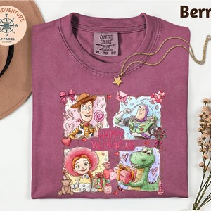 a purple t-shirt with a graphic design of characters from the Toy Story movie, including Woody, Buzz Lightyear, and an anthropomorphic dinosaur. The design also includes a "Happy Valentine's Day" message. The t-shirt is displayed on a white background, along with a hat, a necklace, and some flowers.