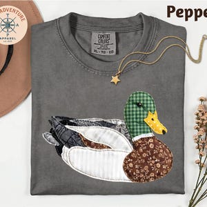 May include: A gray Comfort Colors t-shirt with a duck applique. The duck features a green plaid head, a yellow and white beak, and a brown floral patterned body. A gold star pendant necklace is on the shirt. The word "Pepper" is at the top.