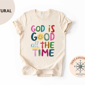 May include: A cream-colored t-shirt with the phrase "GOD IS GOOD all THE TIME" in colorful, handwritten-style letters. A smiling sun graphic is above the word "GOOD". The shirt has a crew neck and short sleeves.