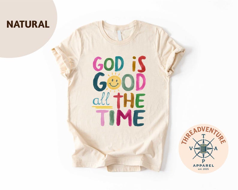 May include: A cream-colored t-shirt with the phrase "GOD IS GOOD all THE TIME" in colorful, handwritten-style letters. A smiling sun graphic is above the word "GOOD". The shirt has a crew neck and short sleeves.