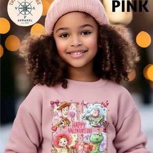 A young girl with curly hair wearing a pink beanie and a pink sweatshirt with a cartoon character design, smiling at the camera.