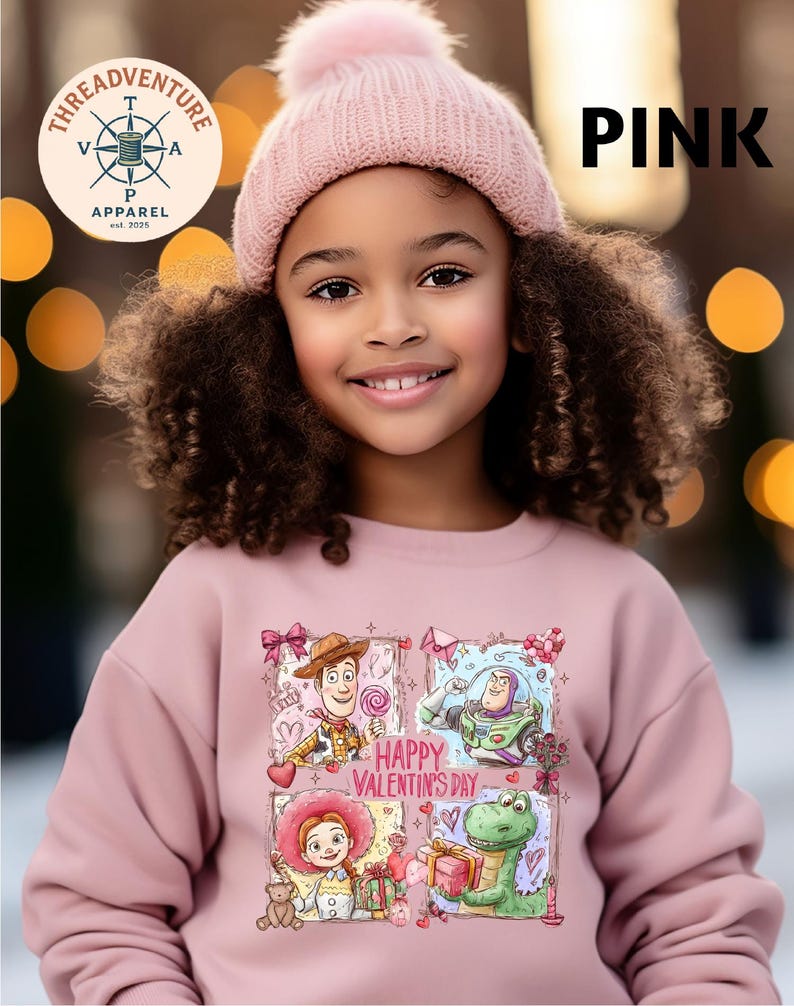 A young girl with curly hair wearing a pink beanie and a pink sweatshirt with a cartoon character design, smiling at the camera.