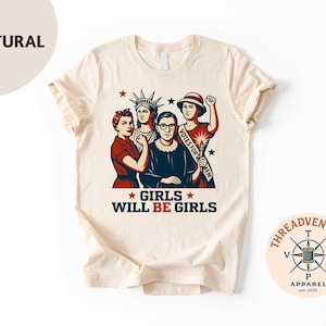 May include: A natural-colored t-shirt featuring a graphic of four women, including Rosie the Riveter and Ruth Bader Ginsburg. The text "GIRLS WILL BE GIRLS" is displayed below the image. The shirt has a crew neck and short sleeves.