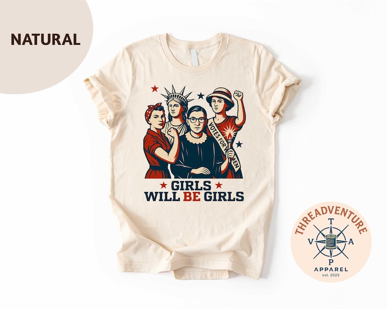 May include: A natural-colored t-shirt featuring a graphic of four women, including Rosie the Riveter and Ruth Bader Ginsburg. The text "GIRLS WILL BE GIRLS" is displayed below the image. The shirt has a crew neck and short sleeves.