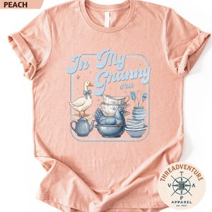 The image is a pink t-shirt with a graphic design featuring a duck surrounded by various kitchen items, such as a teapot, bowls, and a spoon.
