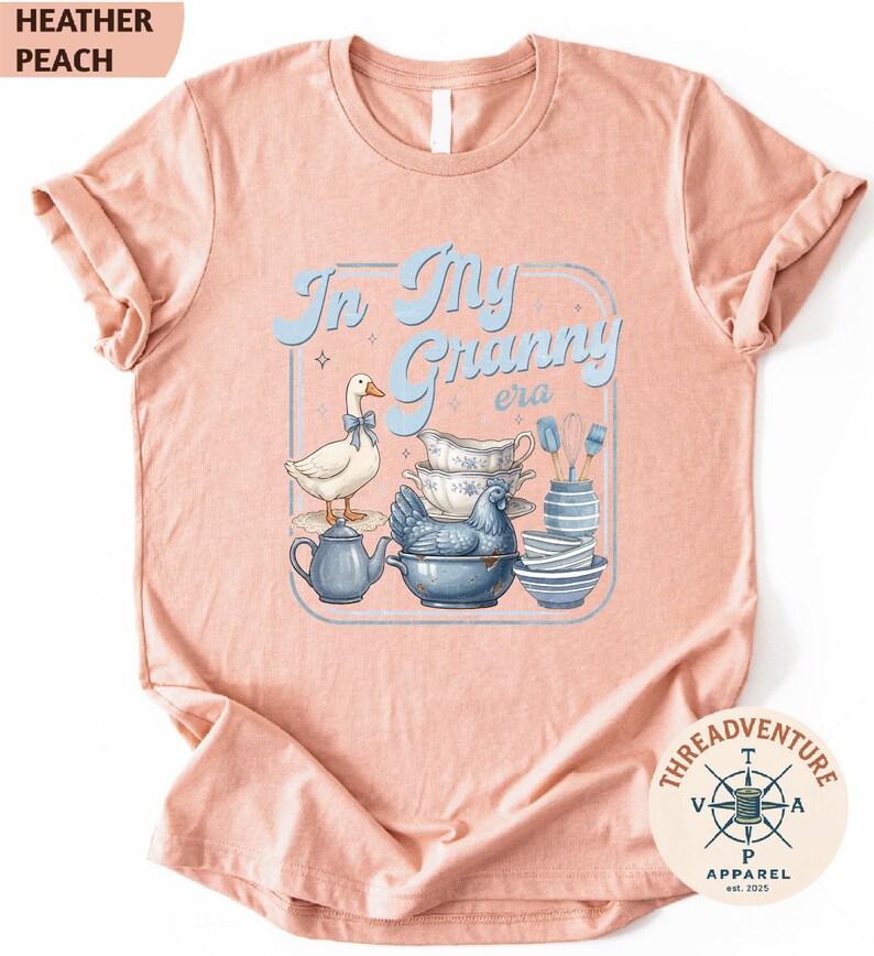 The image is a pink t-shirt with a graphic design featuring a duck surrounded by various kitchen items, such as a teapot, bowls, and a spoon.