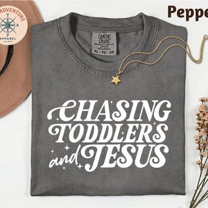 May include: A gray Comfort Colors t-shirt with the text "CHASING TODDLERS and JESUS" in white script. A gold star pendant necklace rests on the shirt. A brown hat and sprig of flowers are in the background.
