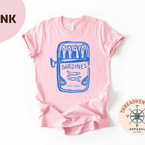 May include: A light pink t-shirt featuring a blue graphic of a sardine can with the word "Sardines" and the text "Wild Caught". The shirt has short sleeves and a crew neck. The Threadventure Apparel logo is in the bottom right corner.