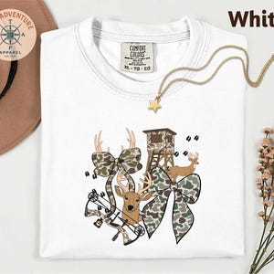 May include: White t-shirt with a deer hunting theme. The shirt features a graphic of a deer, a bow, a hunting blind, and camouflage bows. The text "White" is at the top right. A gold necklace with a star pendant is also visible.