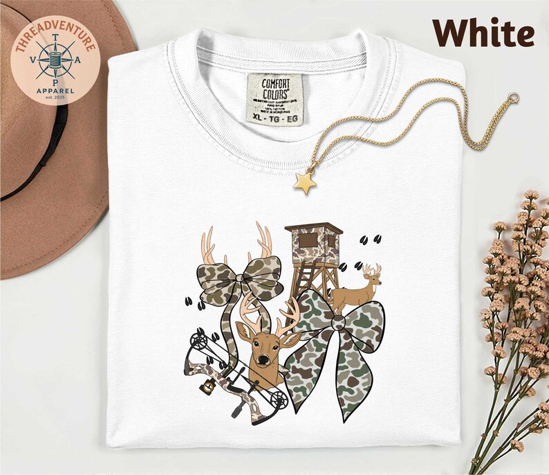 May include: White t-shirt with a deer hunting theme. The shirt features a graphic of a deer, a bow, a hunting blind, and camouflage bows. The text "White" is at the top right. A gold necklace with a star pendant is also visible.