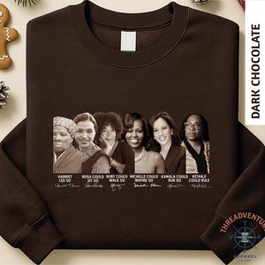 May include: Dark chocolate-colored sweatshirt featuring portraits of historical figures with their signatures and the phrases "Harriet Led So", "Rosa Could Sit So", "Ruby Could Walk So", "Michelle Could Inspire So", "Kamala Could Run So", and "Ketanji Could Rule".