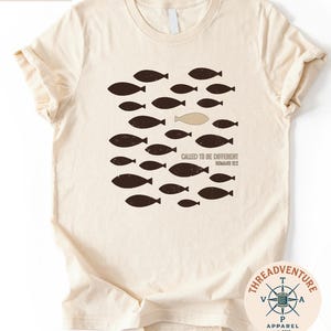 The image is a t-shirt with a graphic design featuring a group of fish, with one fish being different from the rest. The text on the t-shirt reads " Called to be Different" and "Roman 12".
