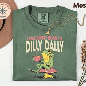 a green t-shirt with a frog sitting on a mushroom and holding a cup, accompanied by a necklace and a hat.