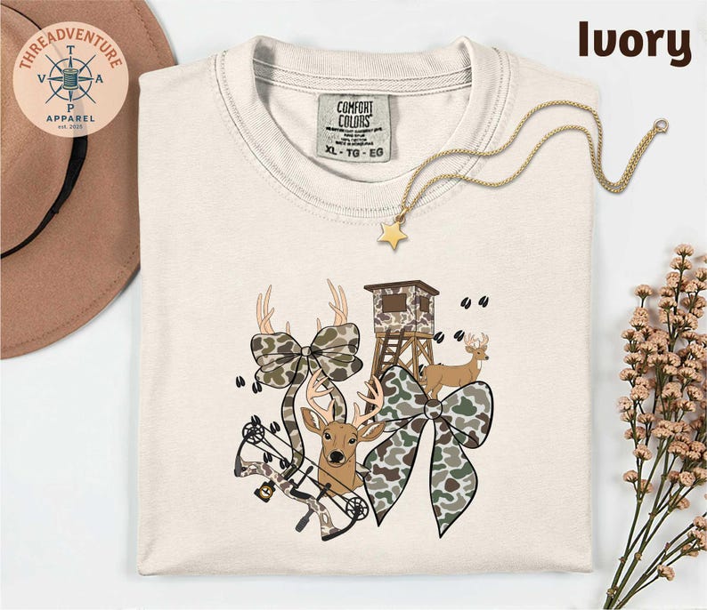 May include: Ivory-colored t-shirt with a hunting-themed graphic. The design includes deer, camouflage bows, a hunting blind, and a bow and arrow. A gold necklace with a star pendant is draped across the shirt.