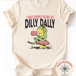 A t-shirt with a cartoon frog sitting on a mushroom and holding a cup of tea. The text on the t-shirt reads "I WAS SIMPLY BORN TO DILLY DALLY".