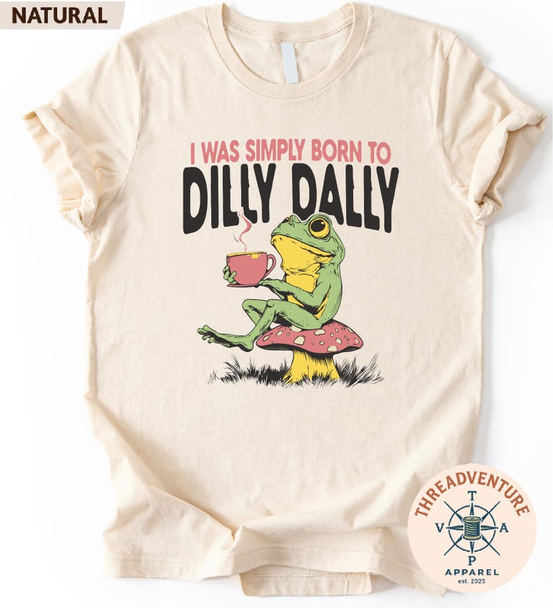 A t-shirt with a cartoon frog sitting on a mushroom and holding a cup of tea. The text on the t-shirt reads "I WAS SIMPLY BORN TO DILLY DALLY".