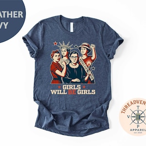 May include: Heather navy t-shirt featuring a graphic of four women, including the Statue of Liberty and Ruth Bader Ginsburg. The text "GIRLS WILL BE GIRLS" is displayed in red and white. The shirt has a crew neck and short sleeves.