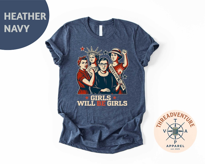 May include: Heather navy t-shirt featuring a graphic of four women, including the Statue of Liberty and Ruth Bader Ginsburg. The text "GIRLS WILL BE GIRLS" is displayed in red and white. The shirt has a crew neck and short sleeves.