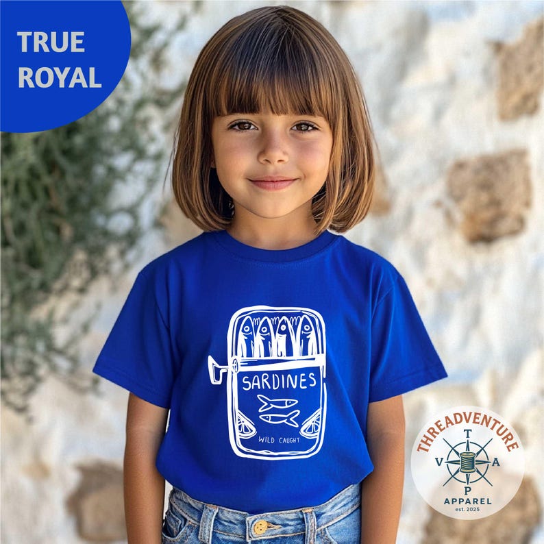 May include: A royal blue t-shirt featuring a white graphic of a sardine can with the word "SARDINES" and "WILD CAUGHT" printed on it. The shirt is worn by a child. The brand logo is visible on the bottom right.