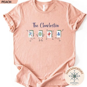 A pink t-shirt with a graphic design featuring four colorful characters, each representing a different element of Charleston, such as a jade, a red and white dragon, a blue and white phoenix, and a green and white kite.