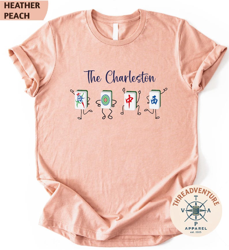 A pink t-shirt with a graphic design featuring four colorful characters, each representing a different element of Charleston, such as a jade, a red and white dragon, a blue and white phoenix, and a green and white kite.