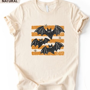 May include: A natural-colored t-shirt featuring a graphic print of three black bats flying over horizontal orange distressed stripes. Small, light-colored stars are scattered among the bats. The shirt has a crew neckline and short sleeves.