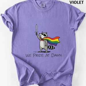 May include: A violet t-shirt featuring a graphic of a raccoon wearing a rainbow cape and holding a sword aloft. Below the raccoon, the text reads WE PRIDE AT DAWN in black capital letters. The shirt has a crew neckline and short sleeves.