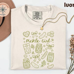 a white t-shirt with a graphic design of various pickle-related items, including jars, a star, and a heart, along with the text "Pickle Girl" printed on it. The t-shirt is laid out on a white background, and there is a hat and some flowers nearby.