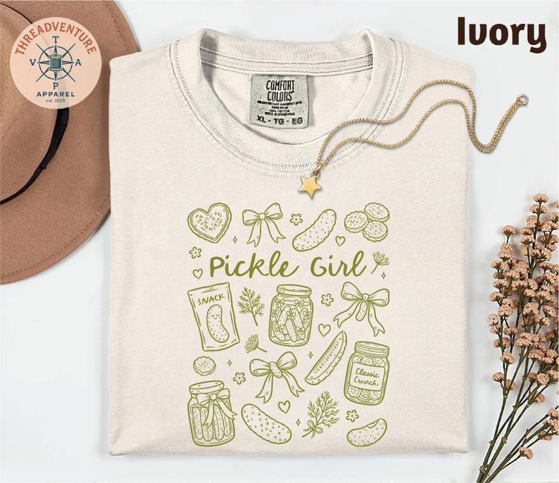 a white t-shirt with a graphic design of various pickle-related items, including jars, a star, and a heart, along with the text "Pickle Girl" printed on it. The t-shirt is laid out on a white background, and there is a hat and some flowers nearby.
