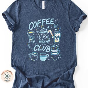 A navy blue t-shirt with a graphic design featuring various coffee-related items such as a coffee pot, coffee cups, and a teapot, along with the text "COFFEE CLUB" and other decorative elements.