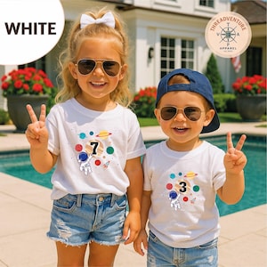 May include: Two children wearing white t-shirts with space-themed graphics, including planets, rockets, and an astronaut. One shirt displays the number 7, the other displays the number 3. Both children are wearing sunglasses and denim shorts.