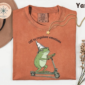 May include: Orange t-shirt with a graphic of a green frog wearing a wizard hat and riding a scooter. The text "Off to regulate emotions" is above the frog. A gold star pendant necklace is on the shirt.