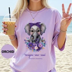 May include: Orchid-colored t-shirt featuring a cute cartoon elephant wearing glasses and a purple bow, surrounded by purple flowers. The text "Forget-me-not Alzheimer's Awareness" is printed below the elephant.
