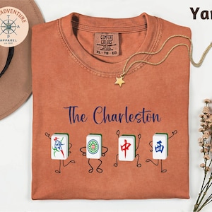 a red t-shirt with a design of four playing cards with different characters on them, along with the text "The Charleston" and a star-shaped charm. The t-shirt is laid out on a white surface, and there is a hat and some flowers in the background.