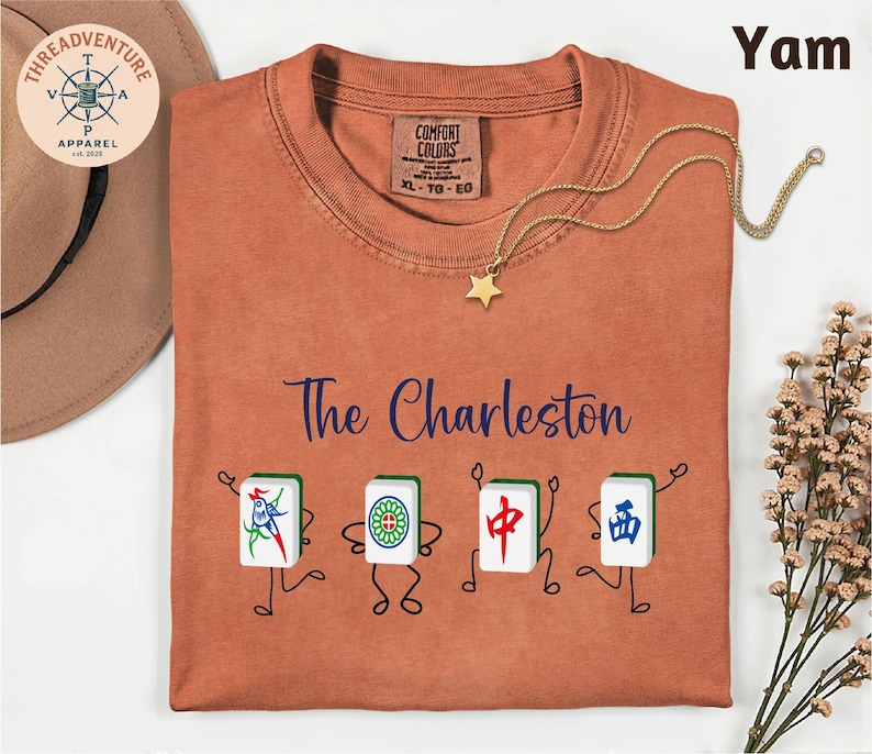 a red t-shirt with a design of four playing cards with different characters on them, along with the text "The Charleston" and a star-shaped charm. The t-shirt is laid out on a white surface, and there is a hat and some flowers in the background.