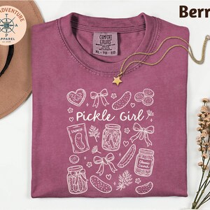 a purple t-shirt with a white design, including various pickle-related items such as jars, a star, and a heart. The t-shirt is laid out on a white surface, with a hat and some flowers nearby.