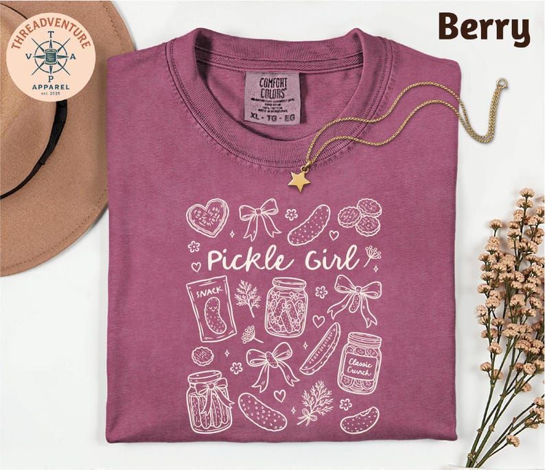 a purple t-shirt with a white design, including various pickle-related items such as jars, a star, and a heart. The t-shirt is laid out on a white surface, with a hat and some flowers nearby.