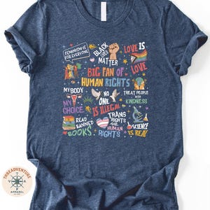 The image is a t-shirt with a collage of various texts and symbols related to human rights and social justice, including phrases like "Black is beautiful", "Love is love", "Big fan of human rights", "My body is my choice", "Treat people with kindness", "Read banned books", "Science is real", and "Trans rights are human rights".
