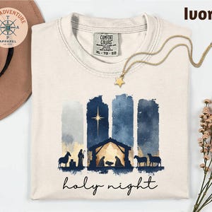 May include: Ivory-colored t-shirt with a watercolor nativity scene design. The design features a star, stable, and silhouettes of figures and animals. The words "holy night" are written in script below the image.