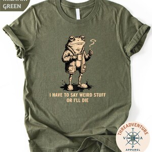 May include: Military green t-shirt featuring a cartoon frog wearing a jacket and holding a cup and a cigarette. The text "I HAVE TO SAY WEIRD STUFF OR I'LL DIE" is printed below the frog. A round logo is in the bottom right corner.
