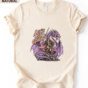 A white t-shirt with a graphic design depicting a woman standing next to a purple dragon. The text on the t-shirt reads "Let's go girls".