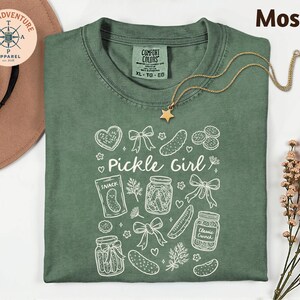 a green t-shirt with a white design, including various pickle-related items such as jars, a star, and a heart. The t-shirt is laid out on a white surface, with a hat and some flowers nearby.
