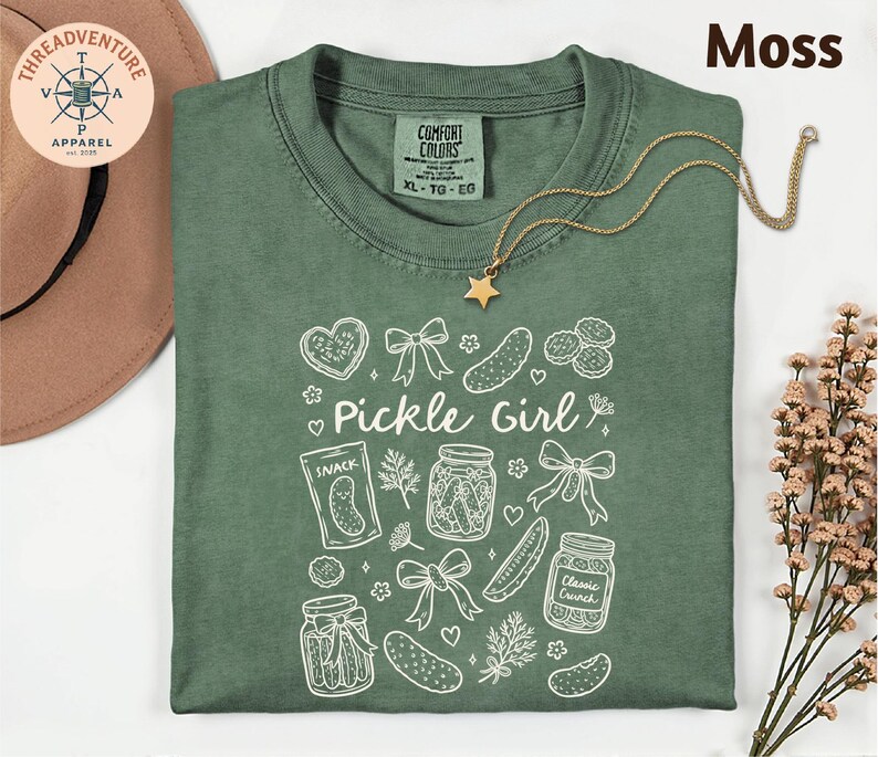 a green t-shirt with a white design, including various pickle-related items such as jars, a star, and a heart. The t-shirt is laid out on a white surface, with a hat and some flowers nearby.