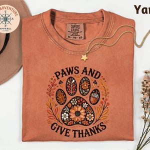 May include: A yam-colored t-shirt with the words "PAWS AND GIVE THANKS" in a circular design featuring a paw print, flowers, and leaves. A gold necklace with a star pendant rests on the shirt. The shirt is from Comfort Colors.