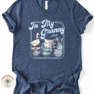 A navy blue t-shirt with a graphic design depicting a white duck surrounded by various kitchen items, including a teapot, bowls, and spoons.